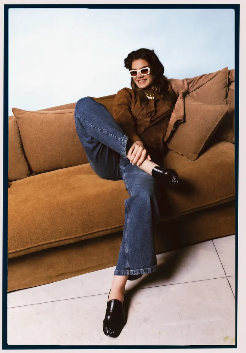 Look L13, Fall Winter 26/27: seduta sul bracciolo, giacca scamosciata marrone e jeans a zampa