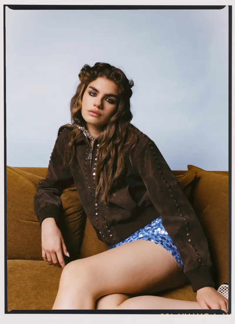 Look L4, Fall Winter 26/27: seduta sul divano, giacca scamosciata marrone con borchie