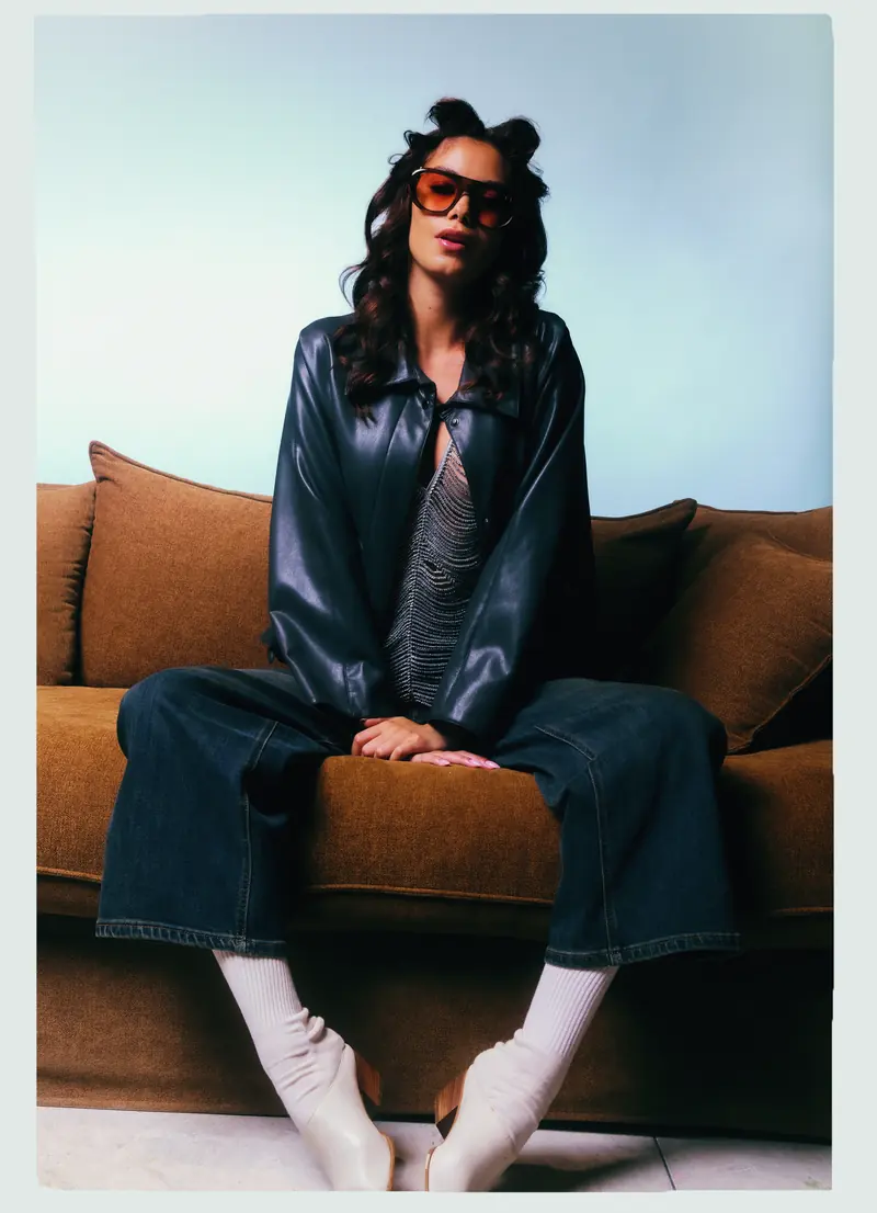 Look L6, Fall Winter 26/27: seduta sul divano, giacca nera lucida e jeans larghi
