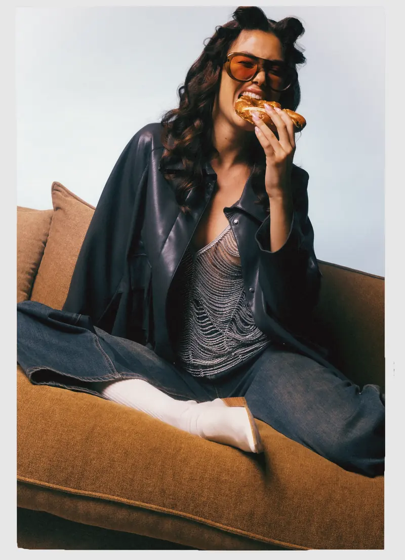 Look L7, Fall Winter 26/27: giacca nera lucida, morde un pretzel sul divano